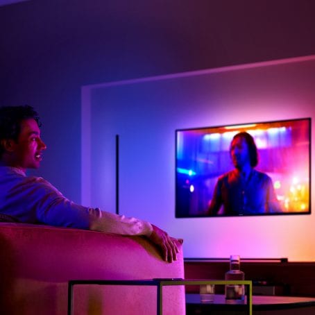 Aan de wand gemonteerde tv met lampen die worden gesynchroniseerd met de inhoud op het scherm, terwijl een man tv kijkt vanaf de bank.